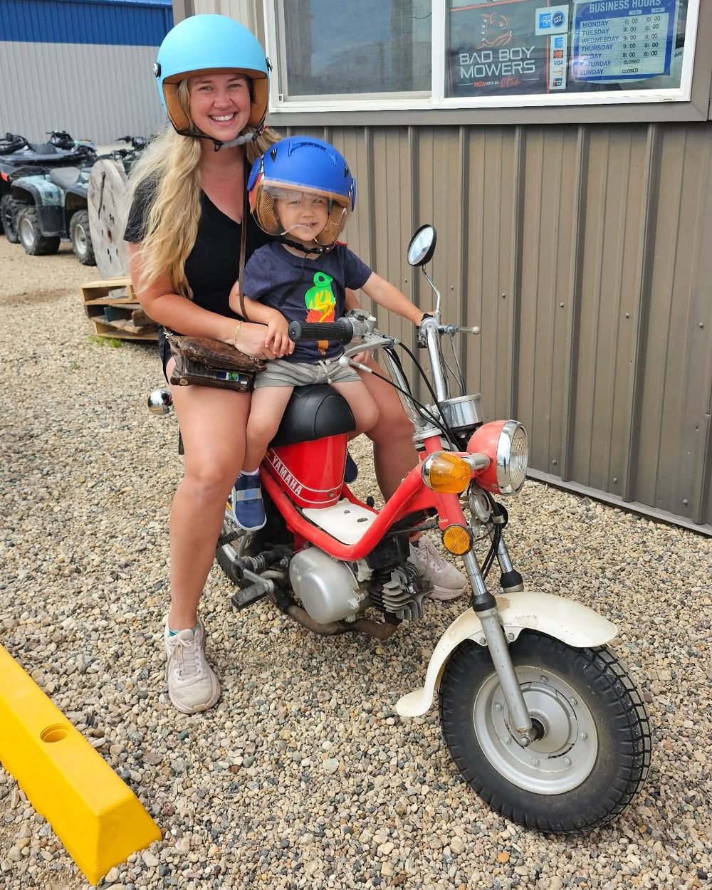 A smiling woman and a small child in helmets on a red Yamaha mini bike outside the shop